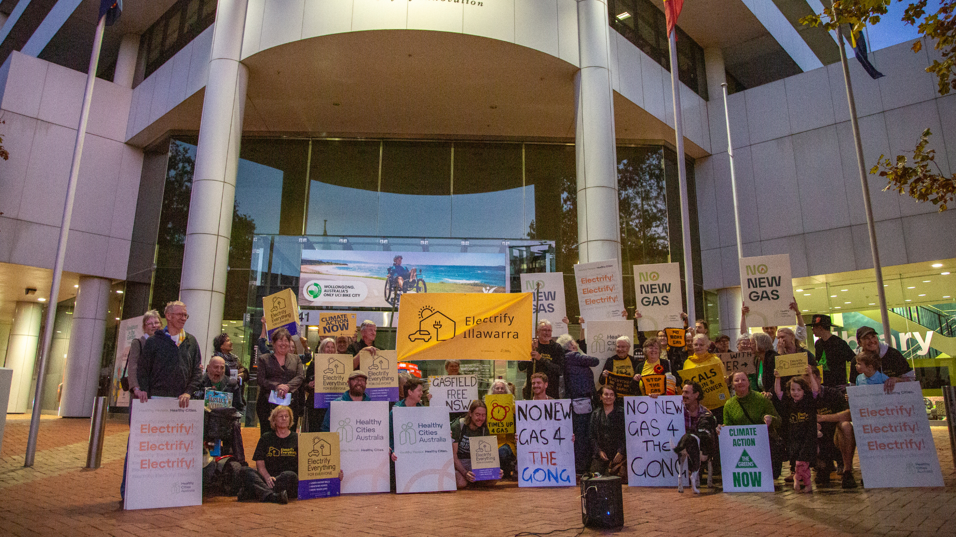 A group of people gathered outside of a building holding signs reading “No New Gas,” “Electrify,” and “No New Gas 4 the Gong.”