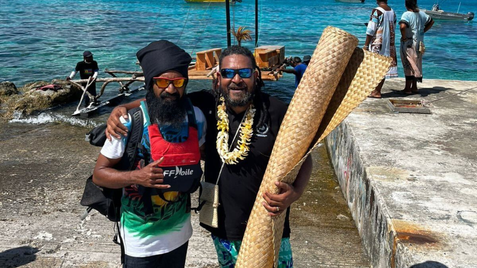 Two men with sunglasses standing together smiling on Lifou Island in New Caledonia. It is sunny. One has his hand wrapped around the shoulders of another and holding a vertical rolled up woven mat and the other man is wearing a small life jacket. They stand on land in front of a woden canoe that is sitting at the shoreline. There is another man standing in the water behind them holding the canoe and two people to the side in the background on a raised concrete platform looking at the canoe. 