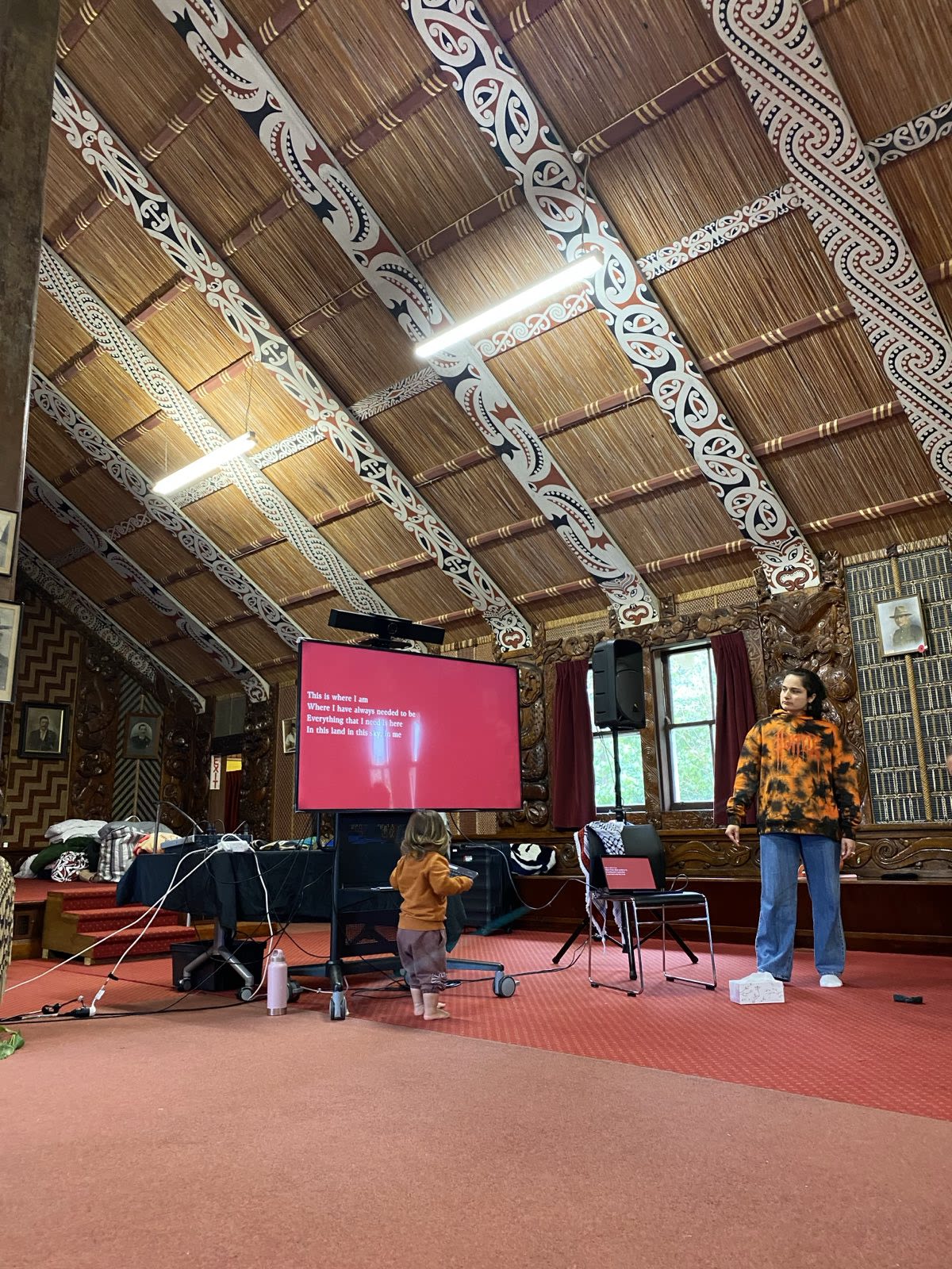 Laniyuk is standing in a marae delivering a workshop. There is a toddler walking towards her.