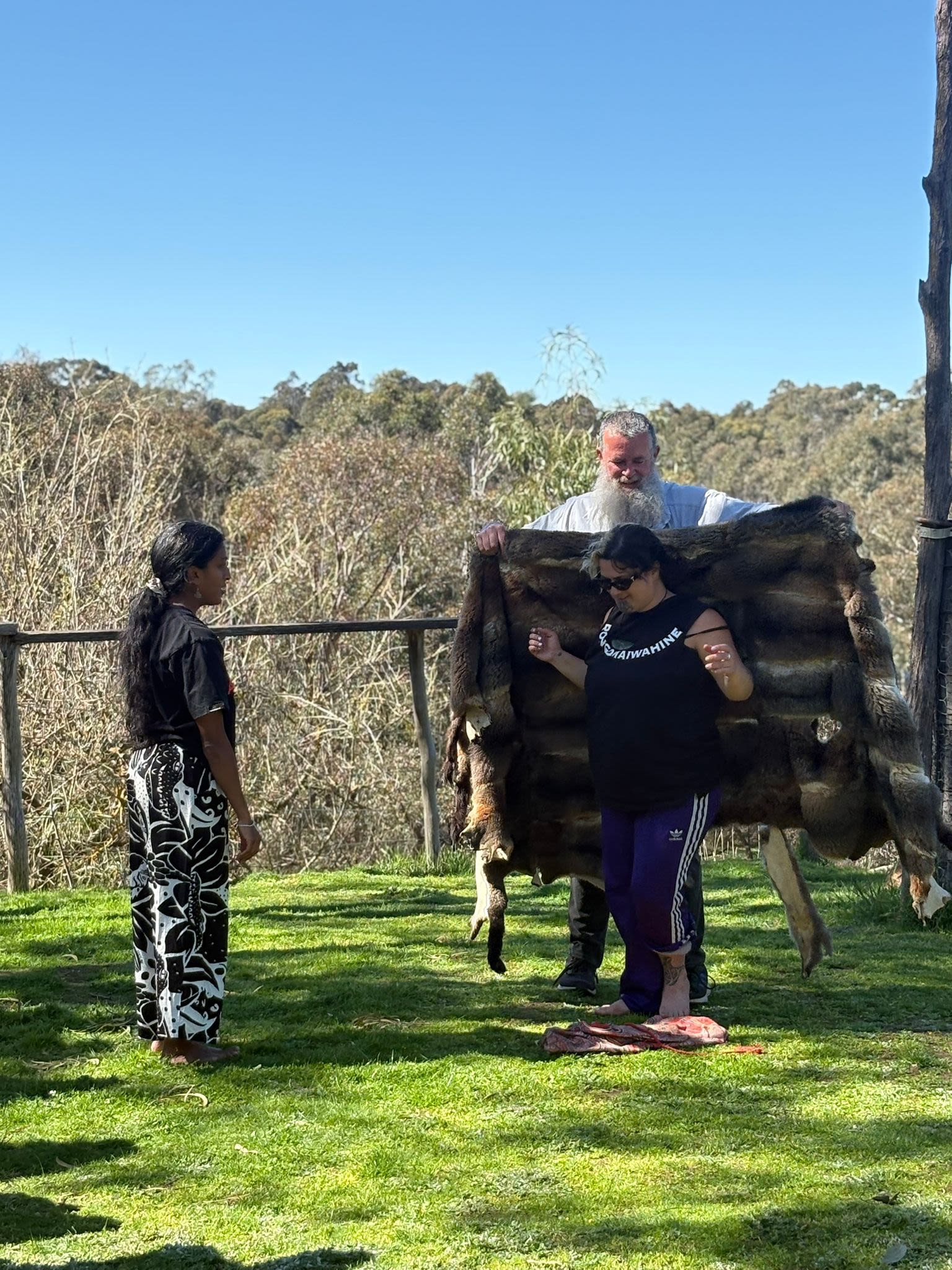 Two people are taking turns to try on the possum cloak that an Elder is holding out for them.