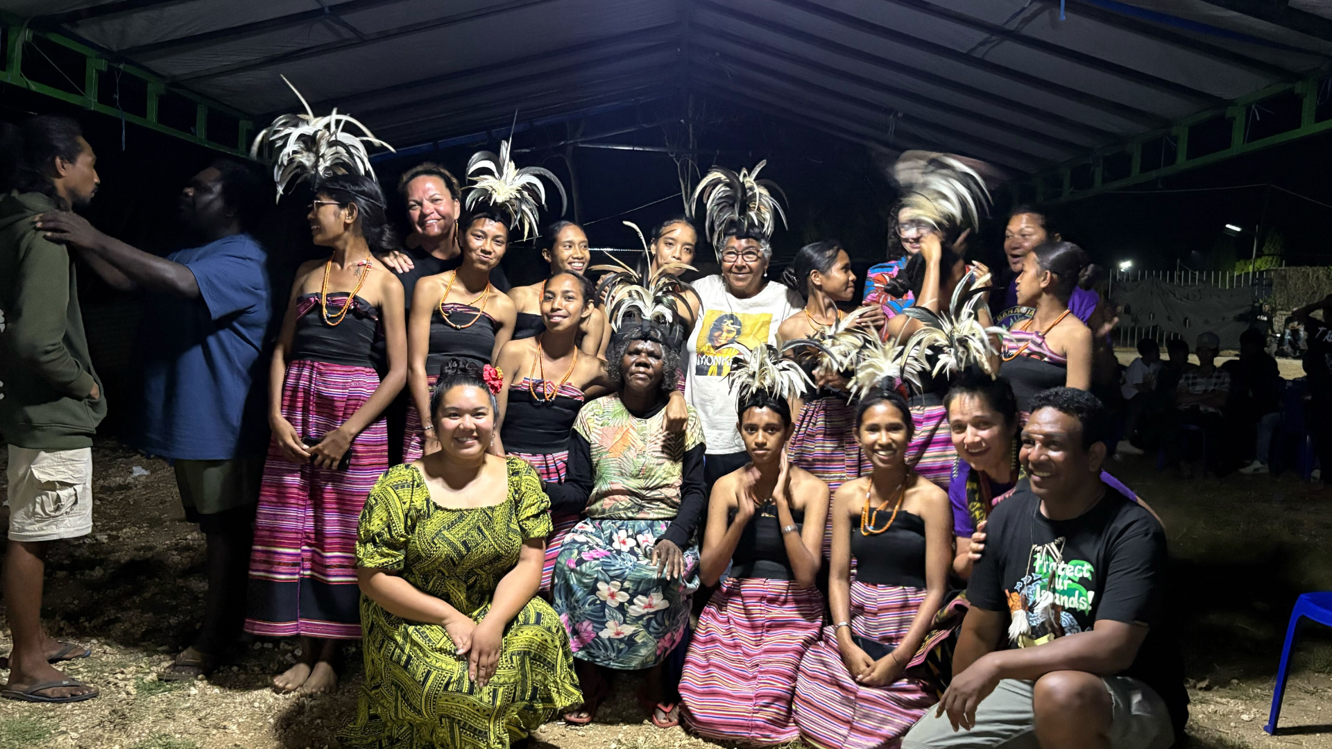 A large group of people pose together under a covered outdoor structure at night. Many wear bright pink and green patterned dresses and skirts, as well as feathered headpieces, and are smiling and looking toward the camera.