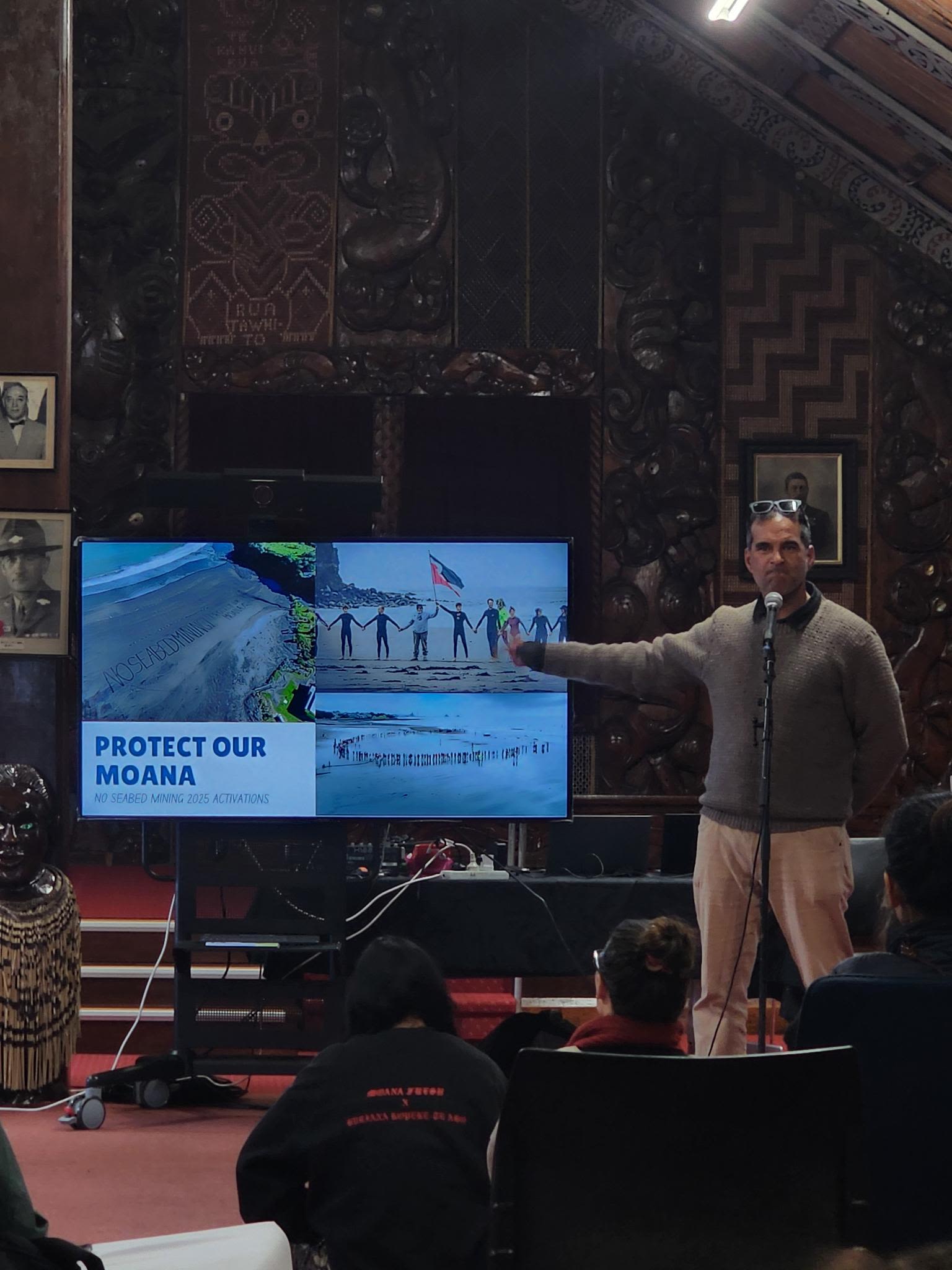 A person stands indoors speaking into a microphone and gesturing toward a presentation on a screen displaying images of a shoreline and people holding hands, overlaid with the text “Protect Our Moana.”