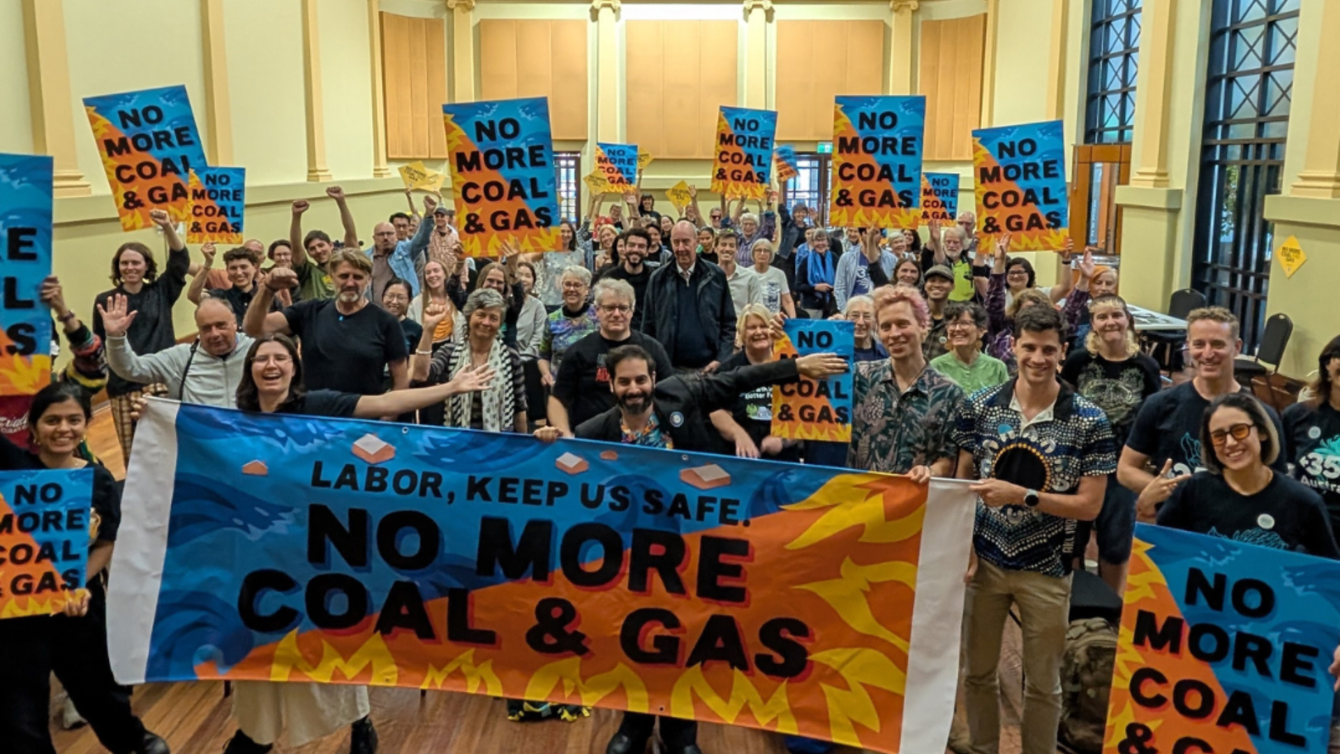 A large group of people gathered indoors holding signs that read “No More Coal & Gas.” People in the front hold a banner that reads “Labor, Keep Us Safe: No More Coal & Gas.”