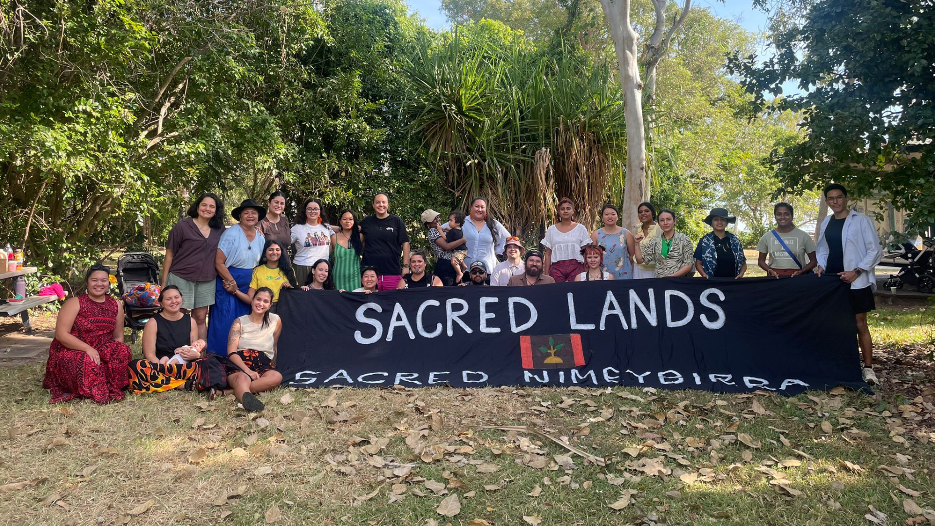 A group of people pose outdoors holding a large banner that reads “Sacred Lands” and “Sacred Nimeybirra”, with a painted image of the Larrakia flag in the centre.