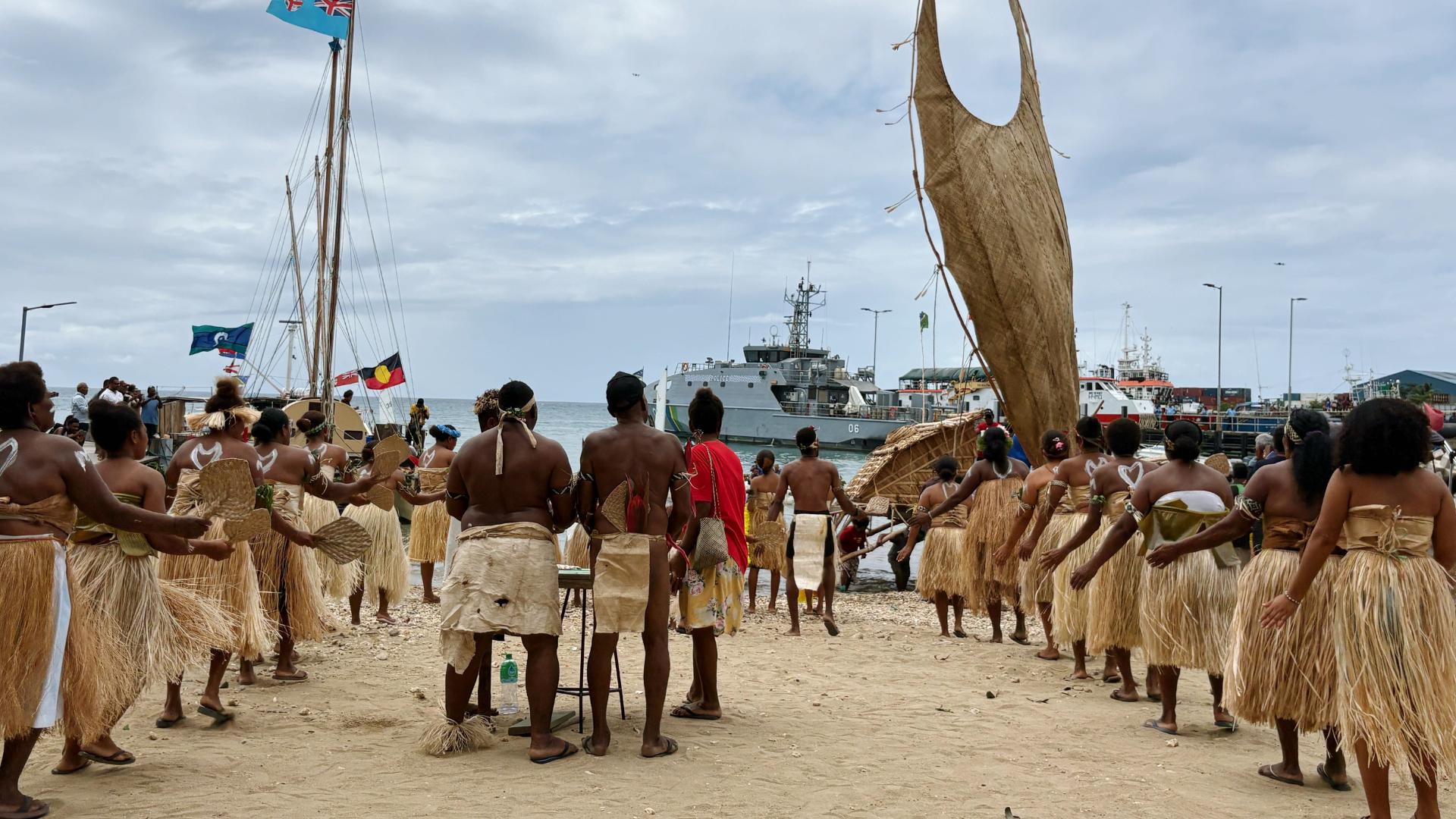 Solomon Islander people dressed in traditional dried grass skirts and dried bark gathered on the shore of a beach in Honiara (capital of Solomon Islands). They are facing the water to acknowledge the arrival of two traditional Canoes - one with a big woven sail mimicking the shape of a man with his hands raised to the sky and the other with tall masts with Pacific flags waving in the wind. Clouds and the horizon cover the top of the image.