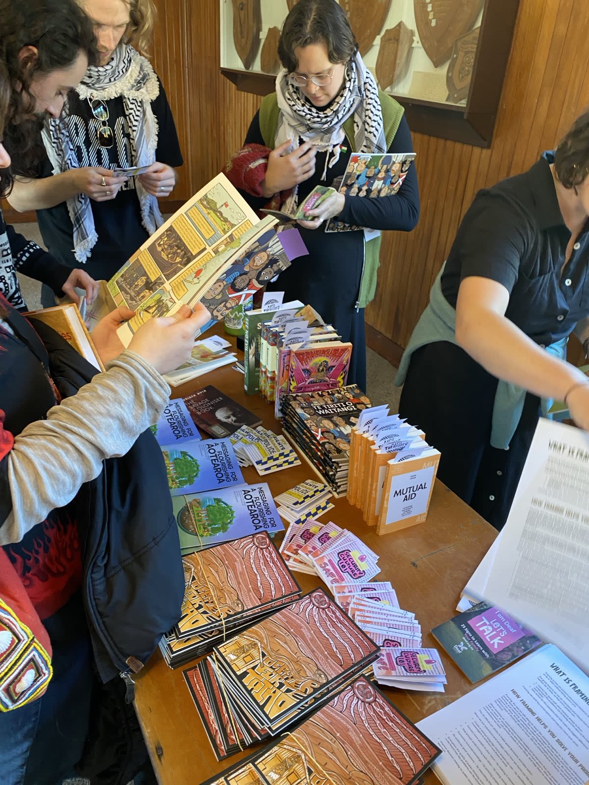People stand around a table covered with pamphlets, zines, and flyers, picking up and reading printed materials.