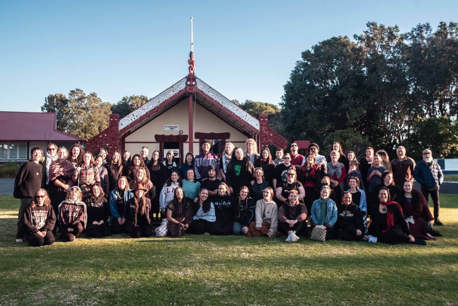 A large group standing in front of a marae with a beautiful pōhutukawa tree in the background