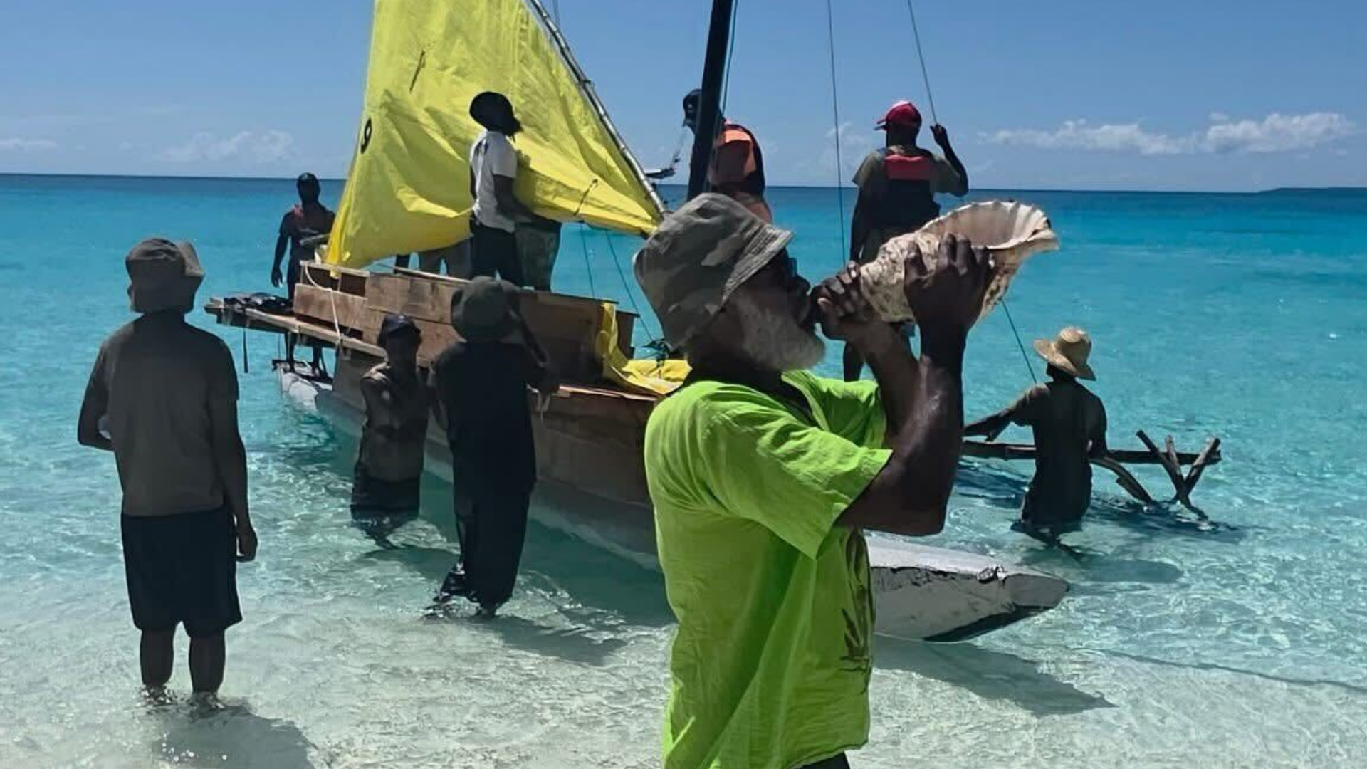 An elder with a hat in the sun infront of the ocean is blowing a trumpet shell as 8 people behind him are assisting each other in getting a canoe with a small sail ready to sail on the water. Some are in the water and some are standing on the canoe. The sail is not yet raised. 