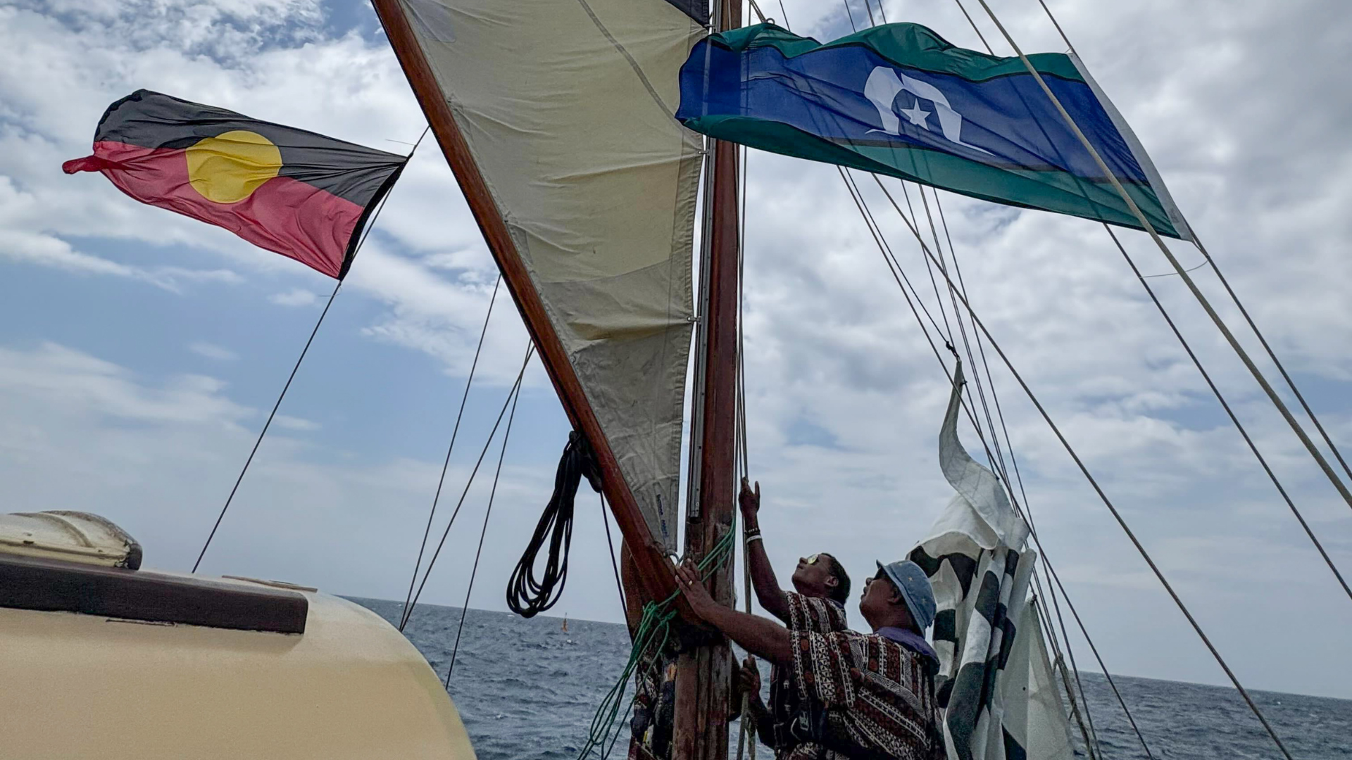 Two people adjust sails on a wooden boat at sea, with the Australian Aboriginal and Torres Strait Islander flags flying from the mast. The ocean and cloudy sky are visible in the background. 