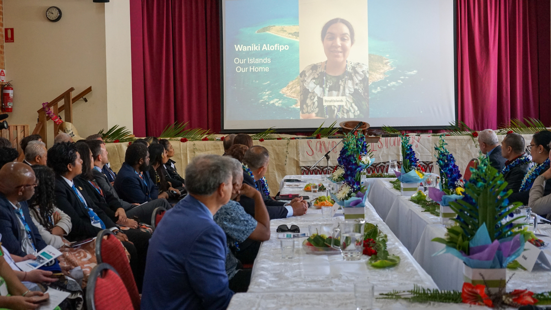 People seated at long tables inside a hall watch a presentation on a large screen showing a speaker and the text “Waniki Alofipo: Our Islands, Our Home.” Decorations and plants line the tables.