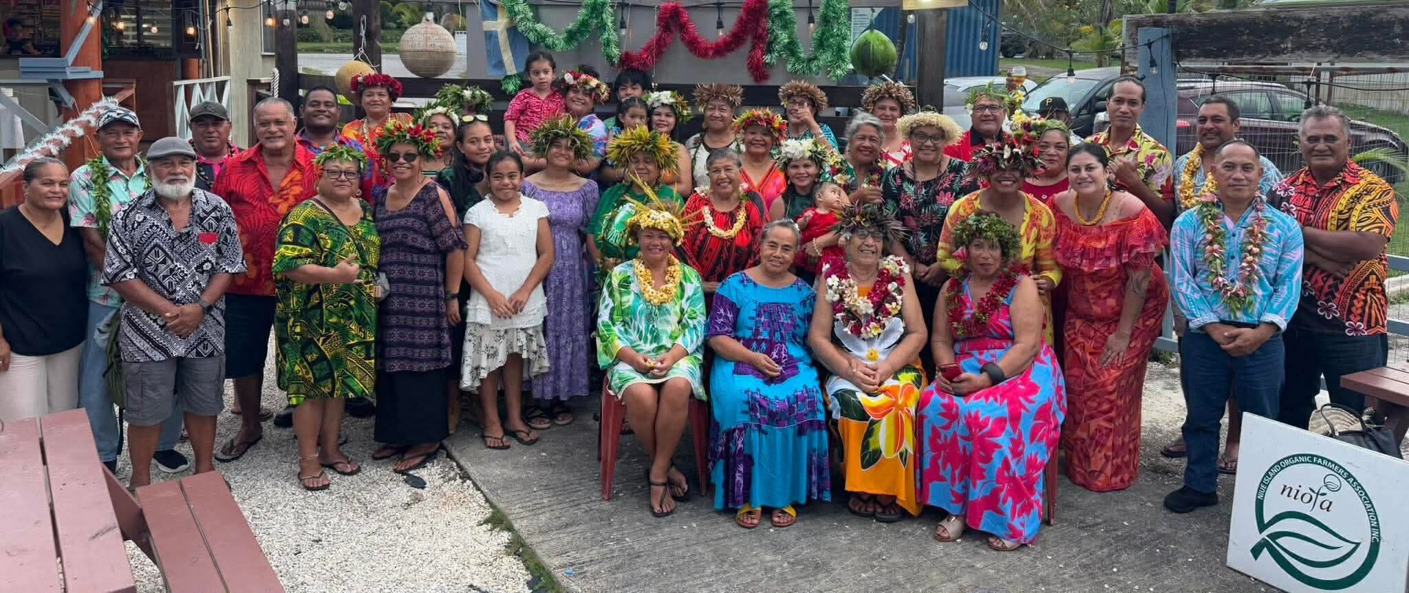 A large group of people gather outdoors for a group photo, many wearing colorful clothing and garlands of flowers and bright green leaves across their heads and necks. 
