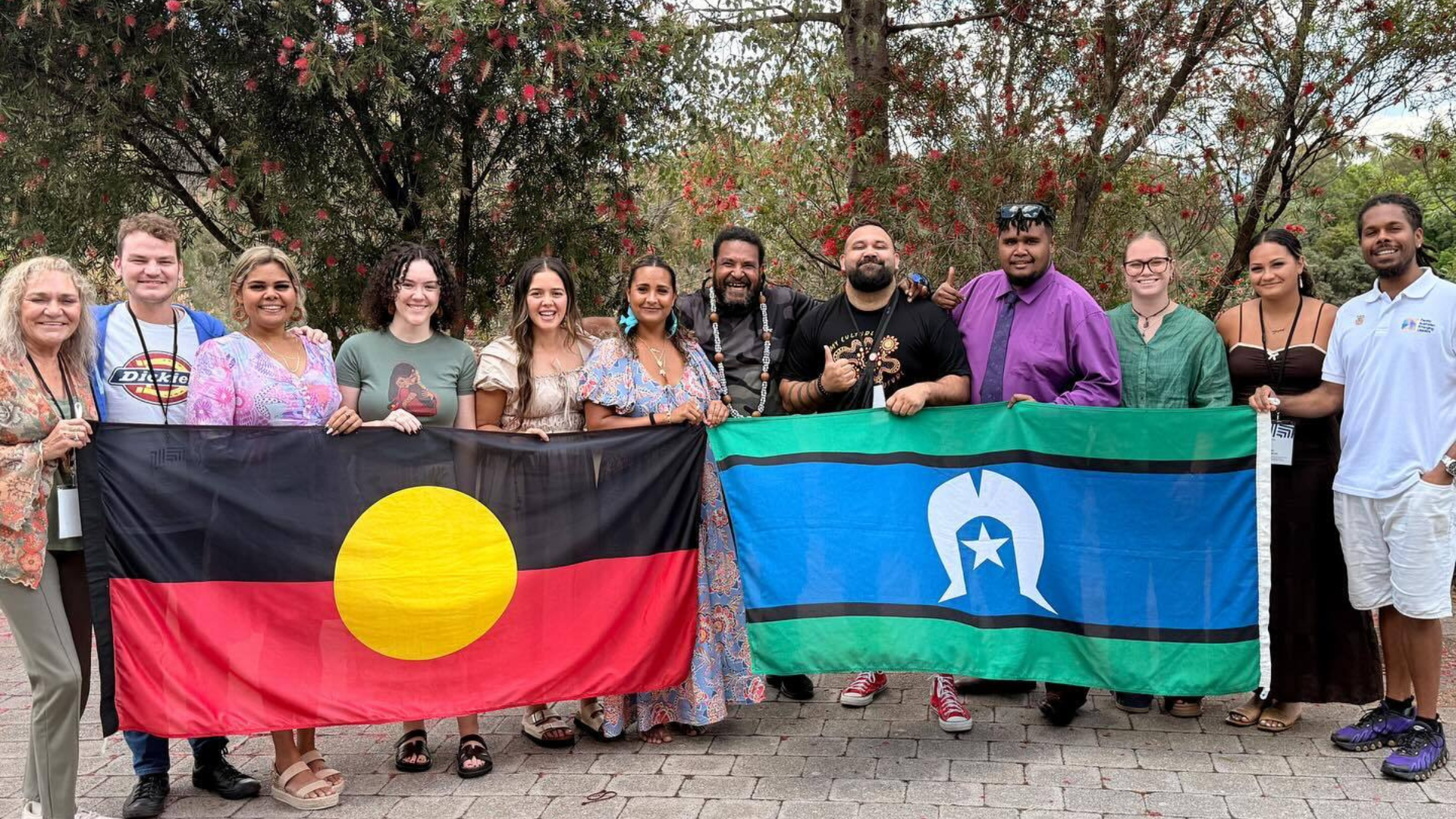 A group of people standing and smiling outdoors in a line, holding the Australian Aboriginal flag and the Torres Strait Islander flag side by side.