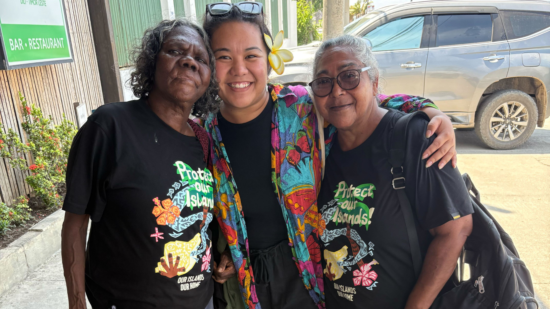 Three people stand close together outside with their arms around each other, smiling at the camera. Two of the people pictured wear matching black shirts printed with the words, “Protect Our Islands”. 