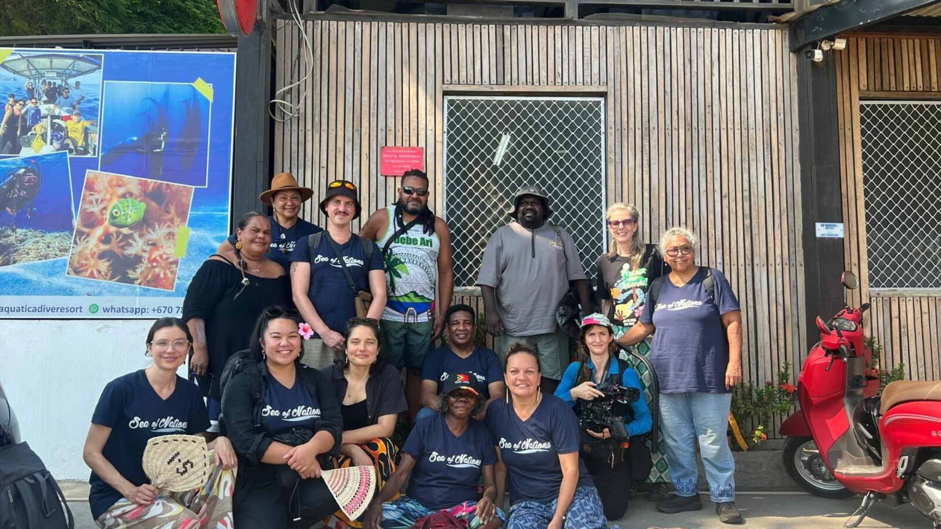 A group of people pose together outdoors in front of a wooden building, smiling at the camera. Many members of the group wear dark blue t-shirts with the words, “Sea of Nations” printed across the front in white. In the background is a presentation featuring photos of marine life. 
