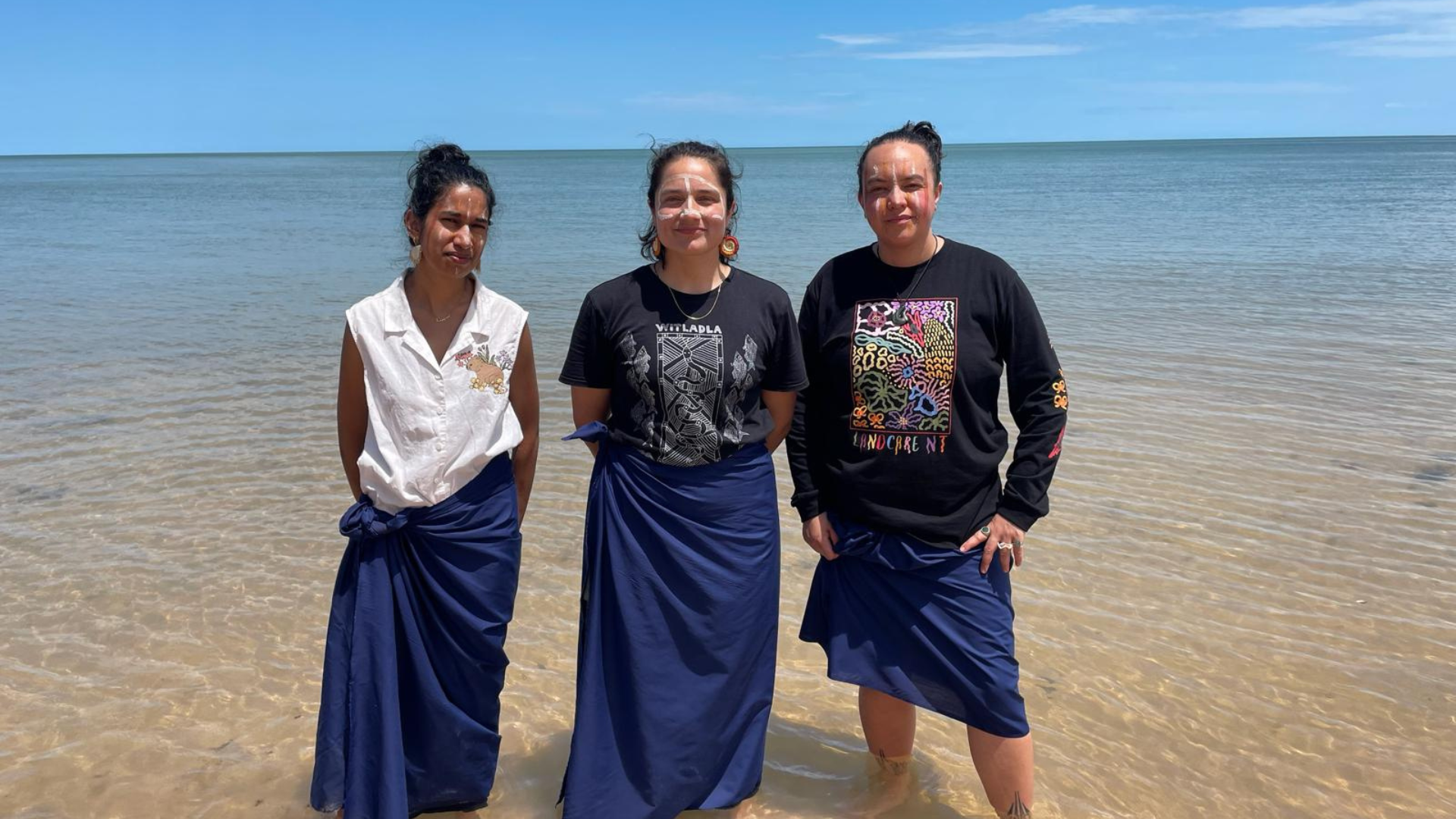 Three people stand in shallow ocean water near the shore, wearing long blue wrap-style garments. It’s a sunny day, and the sea and sky are in the background. 