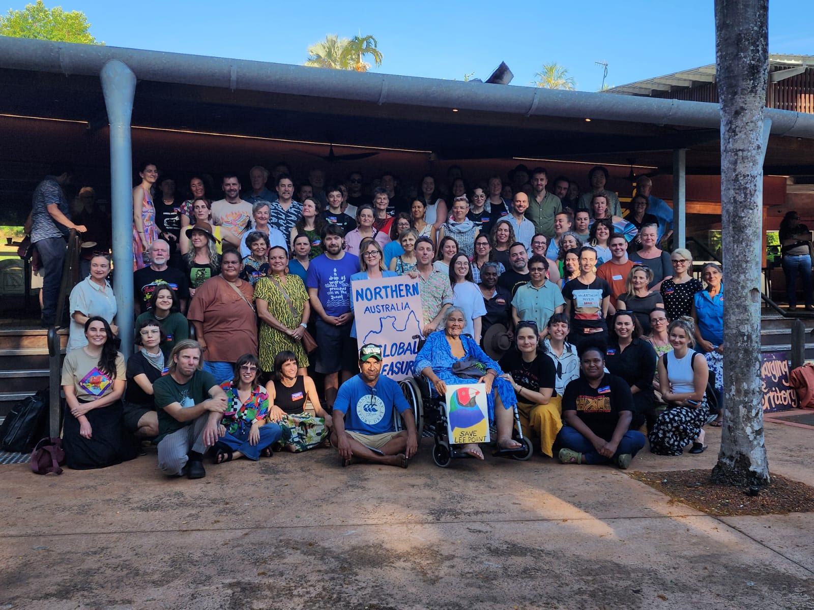 A large group of people pose together outdoors under a covered structure at the Great North Conference in Darwin. Several people hold signs, including one reading “Save Lee Point.”