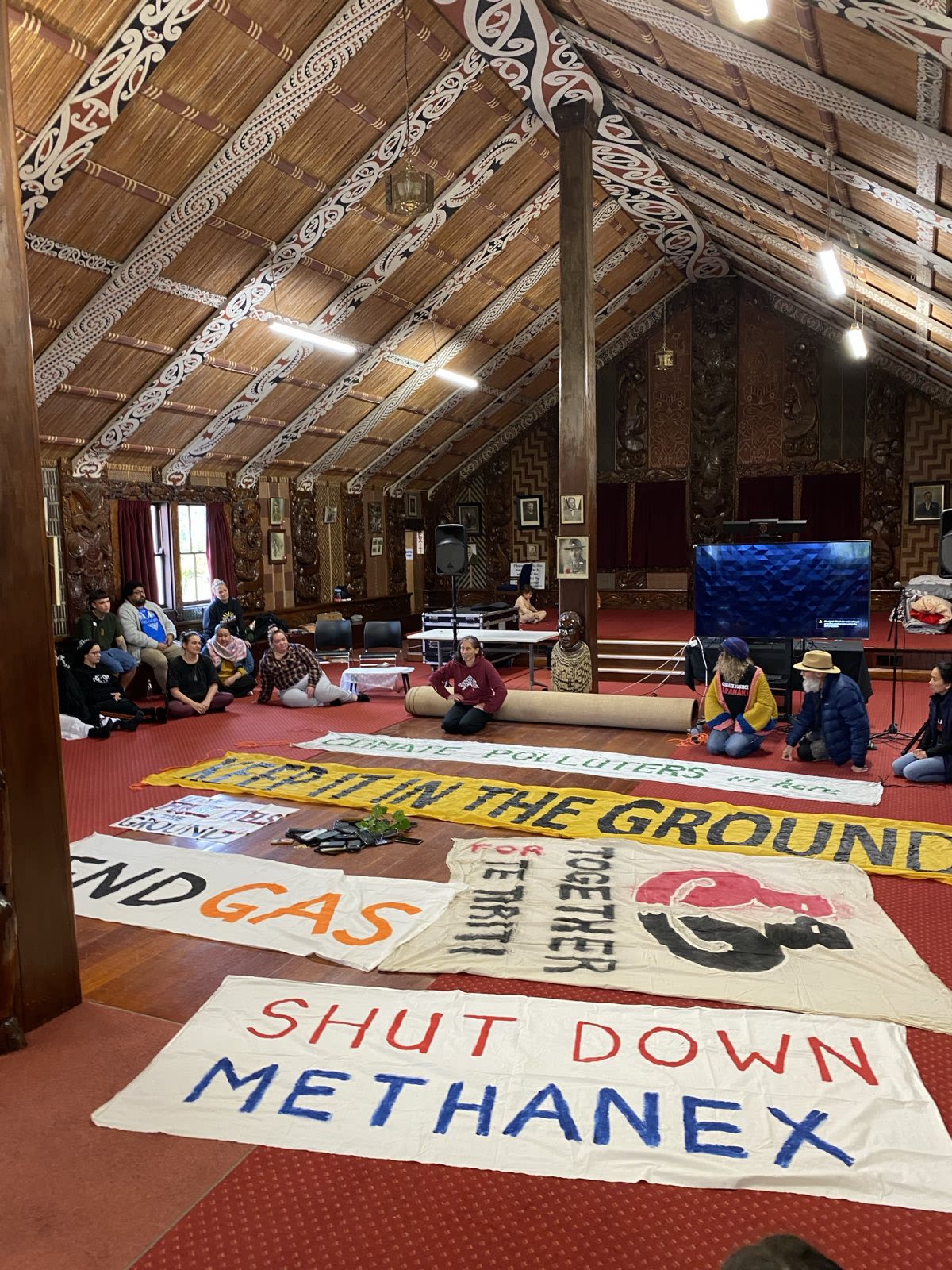 People sit on the floor indoors around large handmade banners laid out on the ground. The room has wooden walls that are carved into various sculptures and patterns throughout the room. One of the banners in the center of the room reads, “Keep it in the Ground”. 