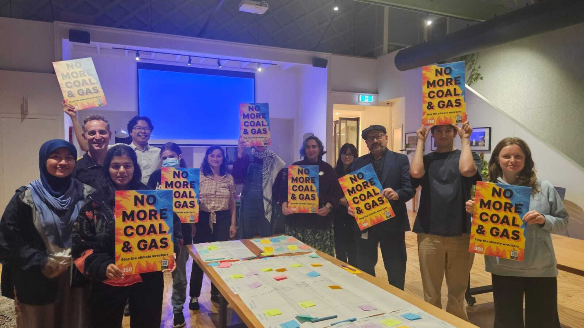 A group of 350 staff and volunteers stand smiling indoors holding signs that read “No More Coal & Gas,”. 