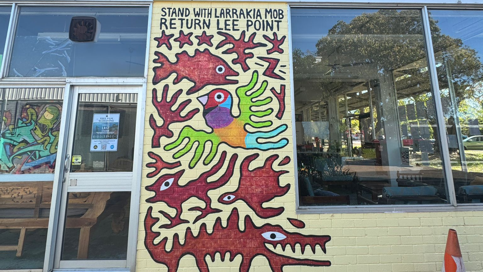 A painted mural on the exterior wall of a building reads “Stand with Larrakia Mob, Return Lee Point.” The mural features stylized animal and plant shapes in red, green, and yellow, with a colourful bird at the centre.
