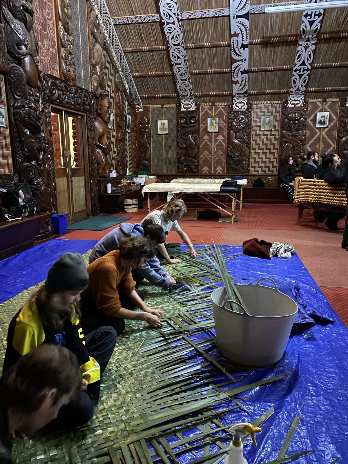 Several people kneel on a tarp indoors, weaving long green leaves together by hand