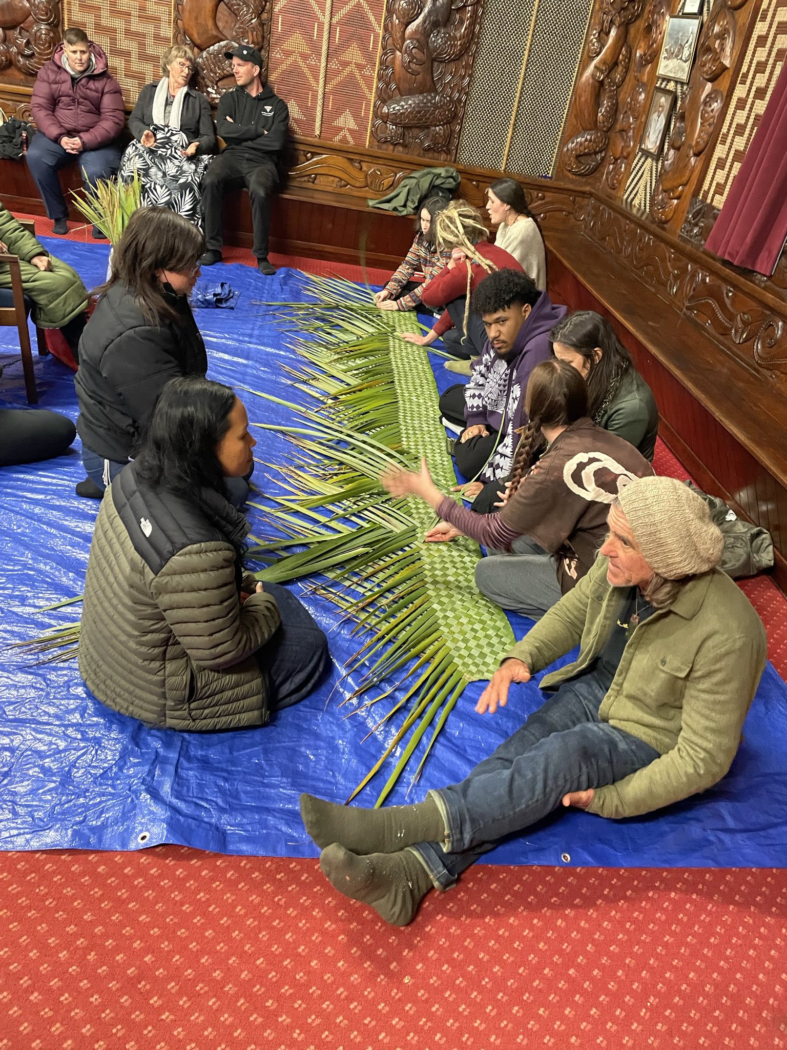 Several people sit on a tarp indoors, weaving long green leaves together by hand, creating a longer woven piece that is spread out across the floor.