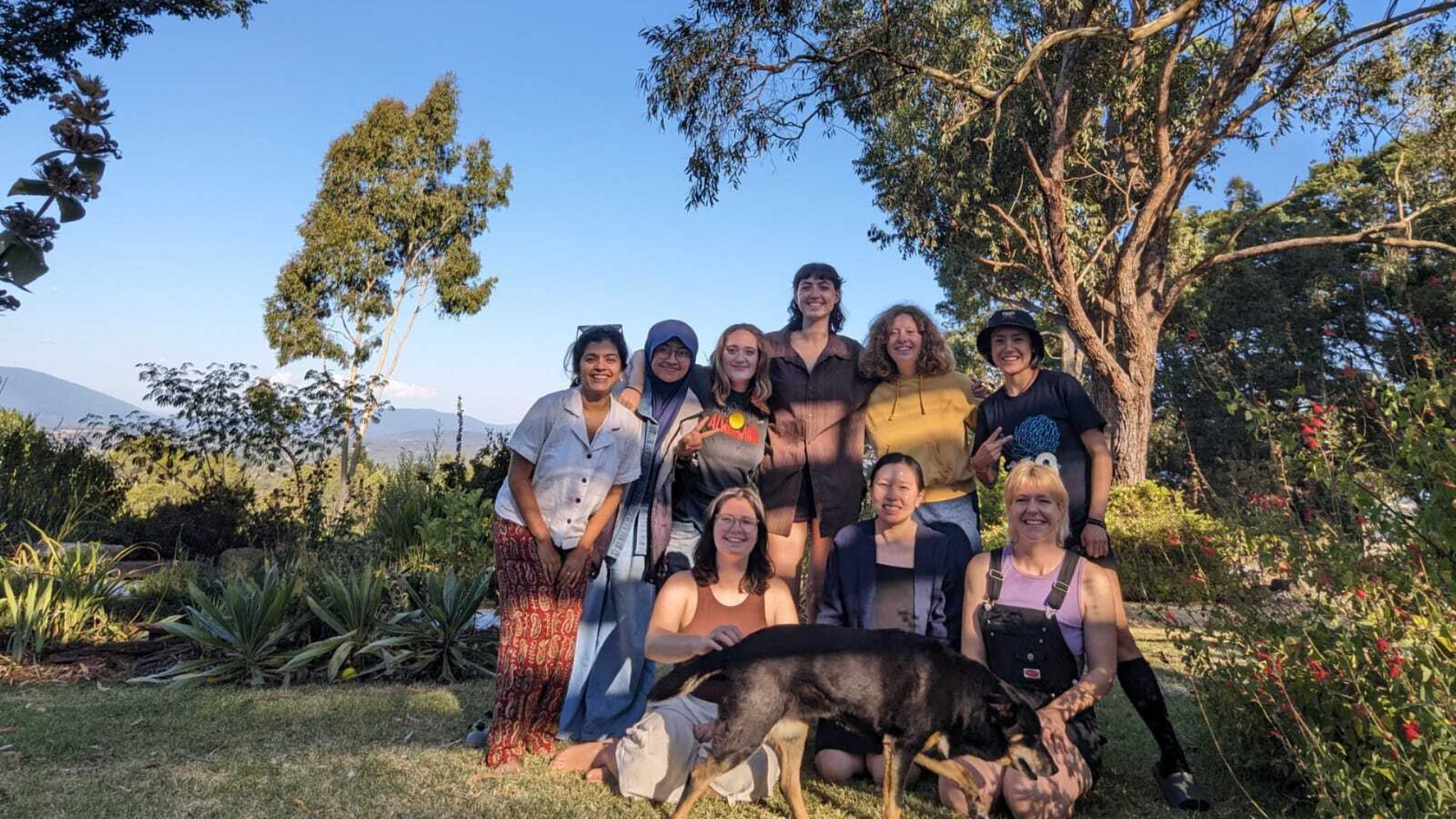 A group of 350 volunteers pose outdoors in front of a lush forest with blue skies and green mountains in the background. The group is standing and kneeling together with a dog in the foreground.
