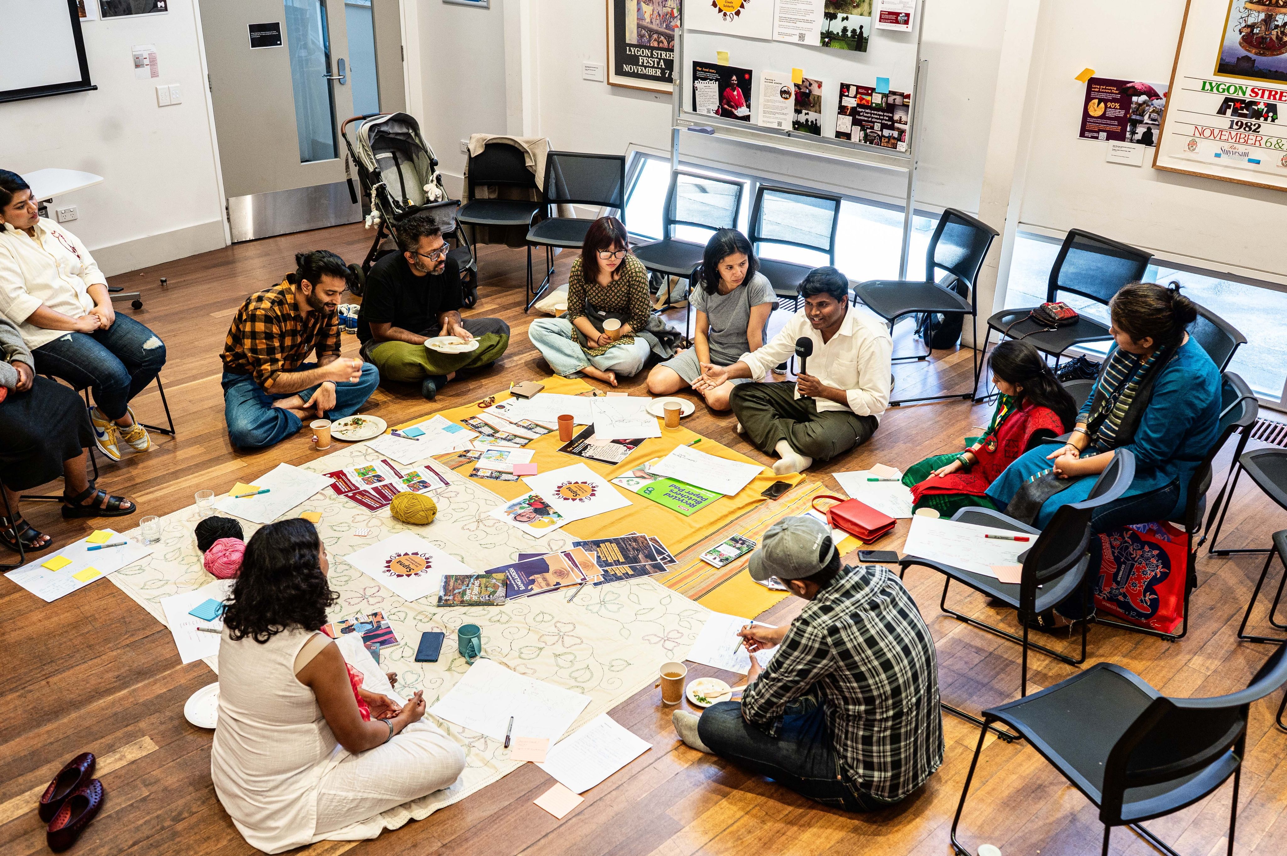 A group of South Asian mostly young people are gathered in a workshop with lots of colourful paper and collage material in front of them 
